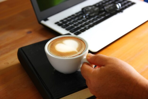 Mano que sostiene el café de la mañana en el tiempo de relajación. Superficie en forma de corazón en la taza de café con leche en el libro negro con material de oficina