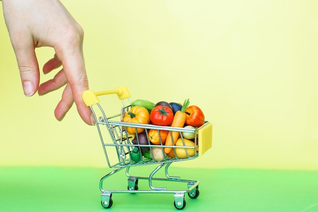 Mano que sostiene el carro de compras de metal con frutas y verduras sobre fondo verde y amarillo