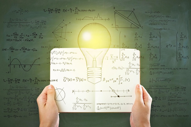 Mano que sostiene el libro abierto con el bosquejo abstracto de la bombilla con fórmulas matemáticas en el fondo de la pared de la pizarra pizarra Concepto de ciencia e innovación de solución de idea de inteligencia