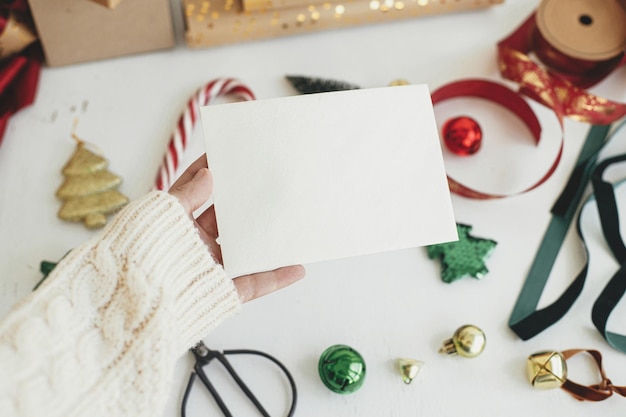 Foto mano que sostiene la maqueta de la tarjeta de navidad tarjeta de felicitación vacía en la mano sobre el fondo de adornos navideños elegantes cintas adornos en la mesa blanca plantilla de postal con espacio para texto