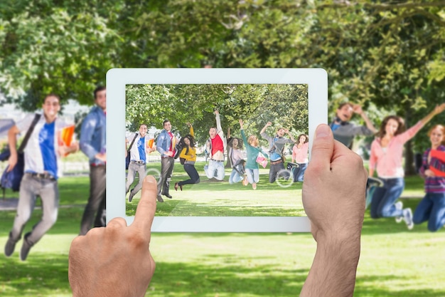 Mano que sostiene la pc de la tableta que muestra a los estudiantes universitarios saltando en el parque