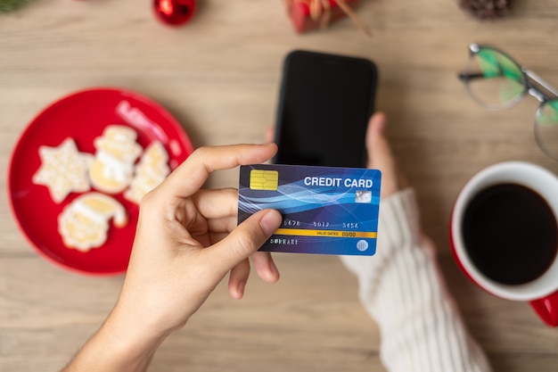 Mano que sostiene la tarjeta de crédito para compras en línea en el teléfono inteligente mientras come galletas caseras de Navidad y café. Feliz víspera de Navidad, diciembre, temporada, venta de viernes negro, año nuevo y concepto de vacaciones