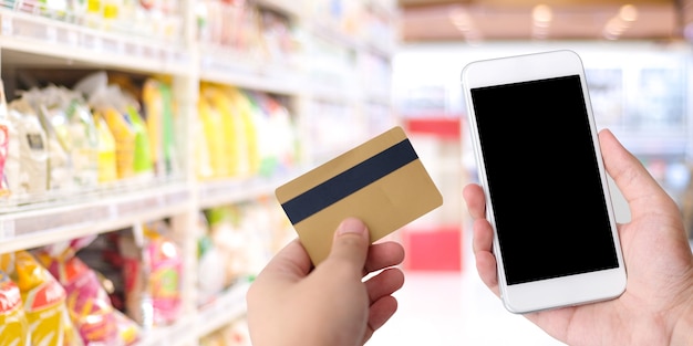 Mano que sostiene la tarjeta de crédito y el teléfono inteligente con pantalla en blanco sobre el supermercado borroso, tienda ba