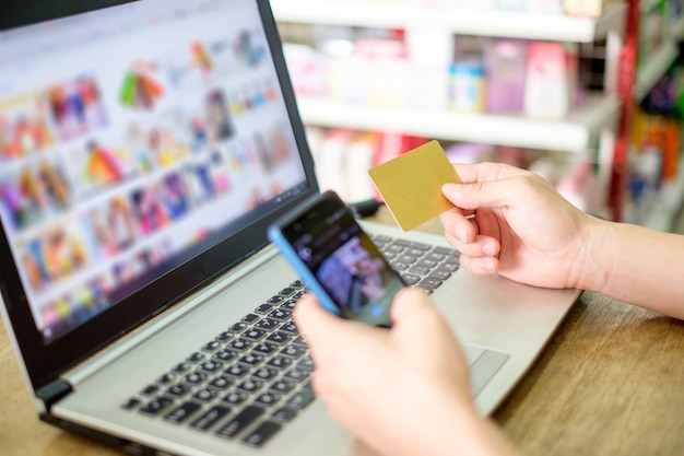 Mano que sostiene la tarjeta de crédito y el uso de una computadora portátil con un teléfono inteligente para realizar compras de pago en línea