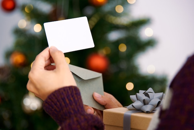 Mano que sostiene la tarjeta de felicitación de navidad de la maqueta y la caja de regalo para el diseño de la invitación en el árbol de navidad