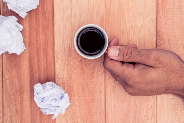 Mano que sostiene la taza de café por papel desmenuzado en el escritorio en la oficina