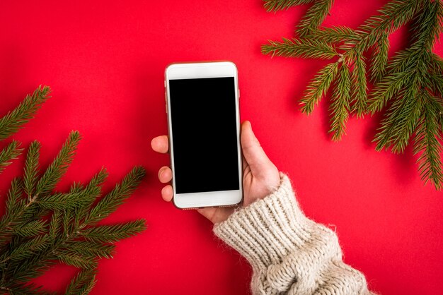 Mano que sostiene el teléfono móvil en rojo con ramas de abeto de Navidad.