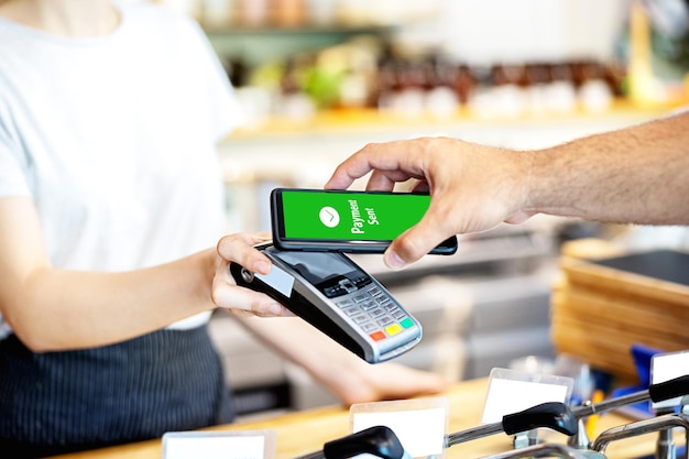Foto mano recortada haciendo pago móvil con lector de tarjetas de crédito sostenido por el propietario en una cafetería