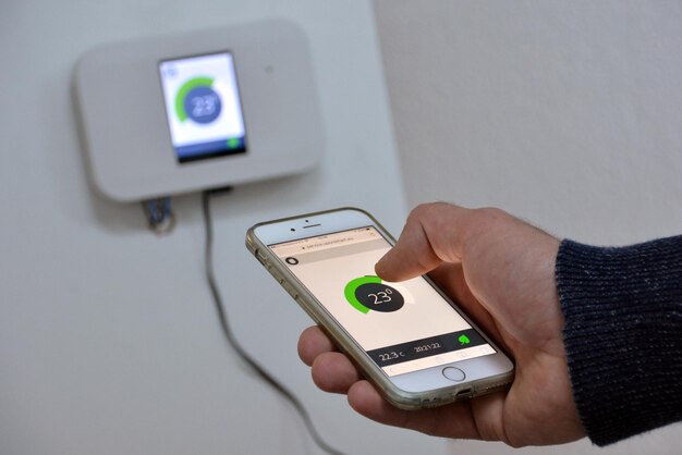 Mano recortada de una persona ajustando el termostato con un teléfono inteligente contra la pared