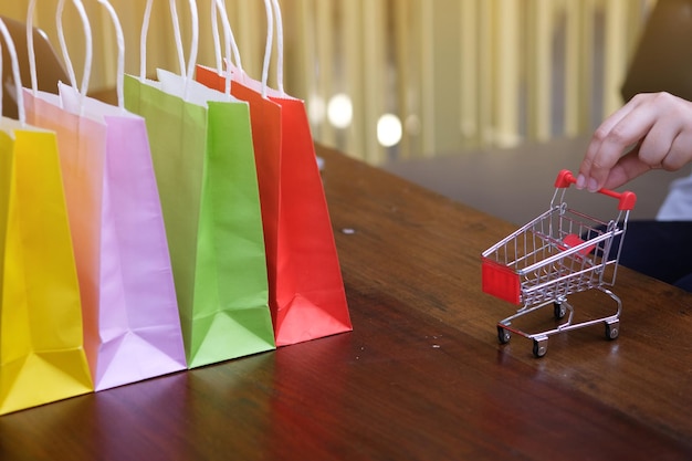 Mano recortada de una persona que sostiene un carrito de compras con bolsas de compras de varios colores en la mesa
