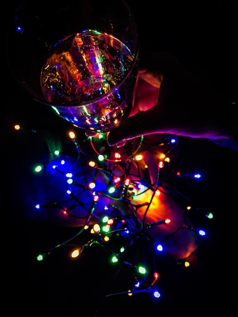 Foto mano recortada sosteniendo un vaso de vino sobre coloridas luces navideñas iluminadas en el cuarto oscuro