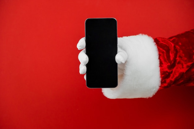 Foto mano de santa claus sosteniendo un teléfono inteligente con una pantalla en blanco.