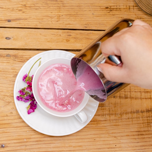 Mano sirviendo un batido de fresa en una taza blanca sobre una mesa de madera