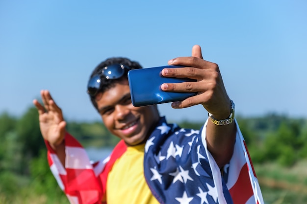 Mano con smartphone de joven cubierto con bandera americana tomando selfie