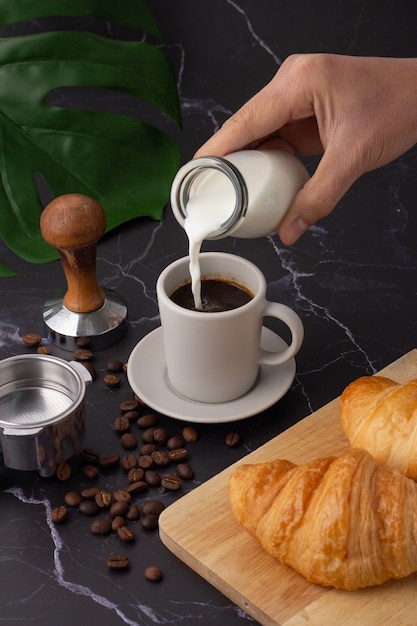 La mano sostenía una botella de leche vertiéndola en una taza de café con leche, croissant en una tabla de cortar, granos de café y un molinillo de café sobre un piso de mármol negro.