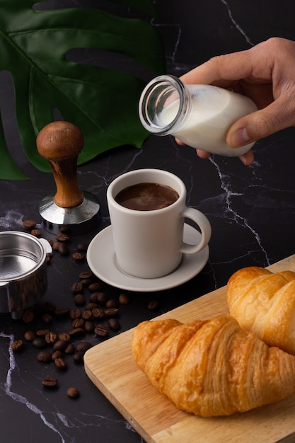 La mano sostenía una botella de leche vertiéndola en una taza de café con leche, croissant en una tabla de cortar, granos de café y un molinillo de café sobre un piso de mármol negro.