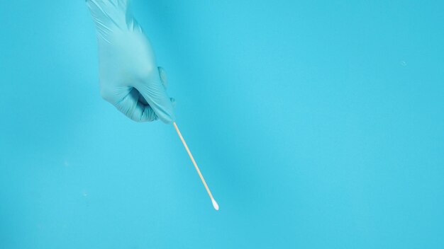 Mano sosteniendo un bastoncillo o bastoncillo de algodón y use guantes médicos azules o guantes de látex sobre fondo azul.