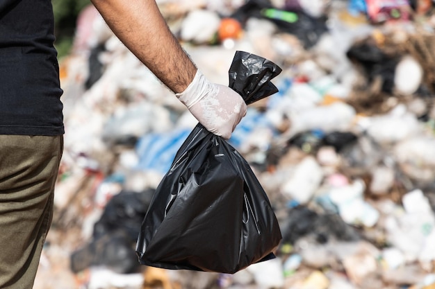 Mano sosteniendo una bolsa de basura negra frente a una gran montaña y un montón de basura. Basuras plásticas de origen industrial y comunitario. Día Mundial del Medio Ambiente.