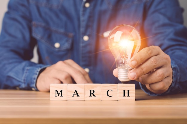 Mano sosteniendo una bombilla con bloques de madera, escriba la palabra MARZO dispuesta en un escritorio de madera con fondo de pared de espacio gris Comience para una nueva idea en el concepto de marzo