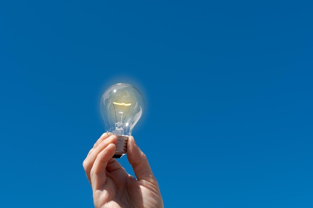 Mano sosteniendo una bombilla eléctrica iluminada con el cielo azul de fondo