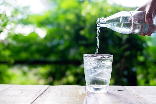 Foto mano sosteniendo una botella de agua vertida en un vaso transparente sobre una mesa de madera fondo verde de la naturaleza