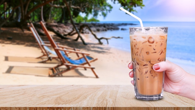 Mano sosteniendo el café helado de vidrio en la mesa de madera con vista al paisaje de playa fondo de naturaleza Café con leche helada