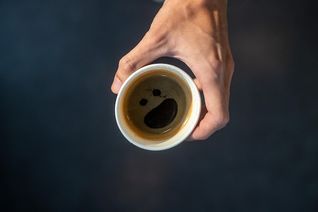 Una mano sosteniendo un café negro en un vaso de papel con una carita sonriente artística en una mesa negra