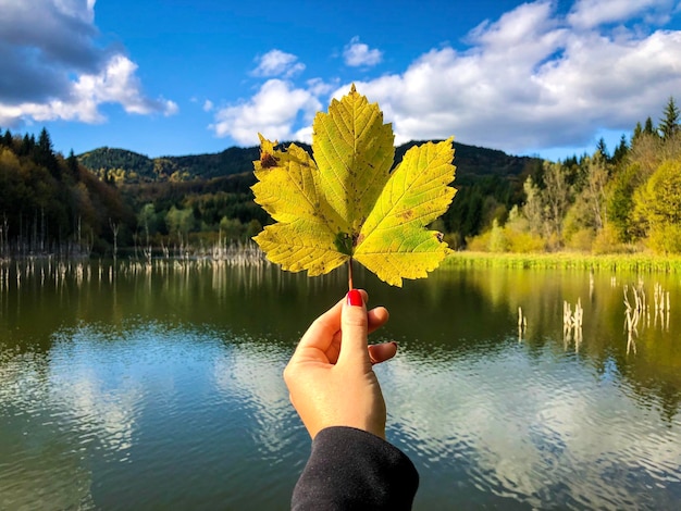 Foto mano sosteniendo hoja amarilla contra el lago y las montañas