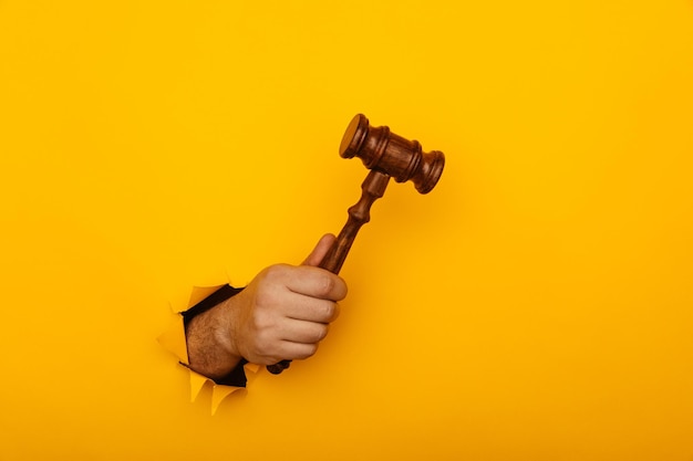 Mano sosteniendo el mazo de un juez a través de la pared de papel amarillo roto Concepto de ley
