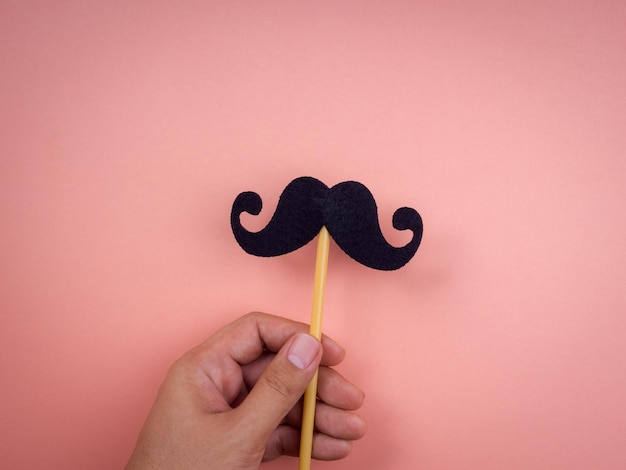 Mano sosteniendo y mostrando bigote palo aislado sobre fondo rosa pastel, estilo minimalista. Feliz día del padre, concepto Movember.