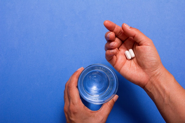 Mano sosteniendo pastillas blancas sobre fondo azul y un vaso de agua. Copie el espacio.