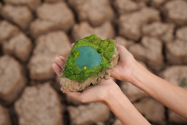 Mano sosteniendo un pedazo de tierra agrietada que tiene un árbol verde y un estanque azul