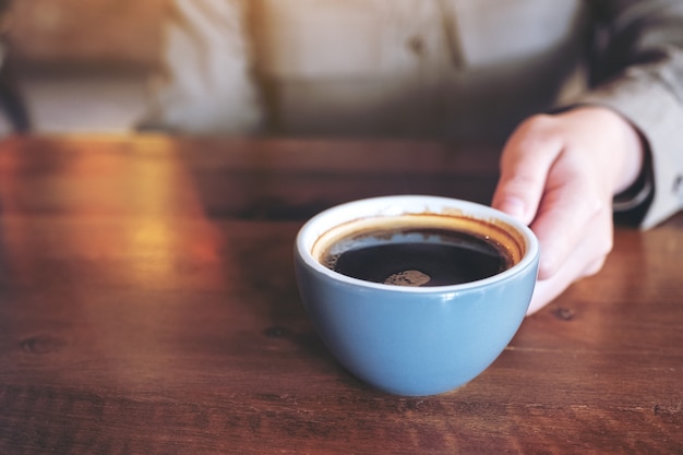 Mano sosteniendo una taza azul de café negro caliente en la mesa de madera en el café