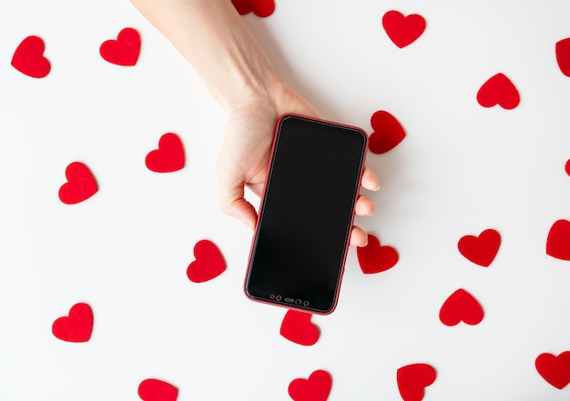 Una mano sosteniendo un teléfono inteligente rodeado de formas de corazón rojo en un concepto blanco del Día de San Valentín