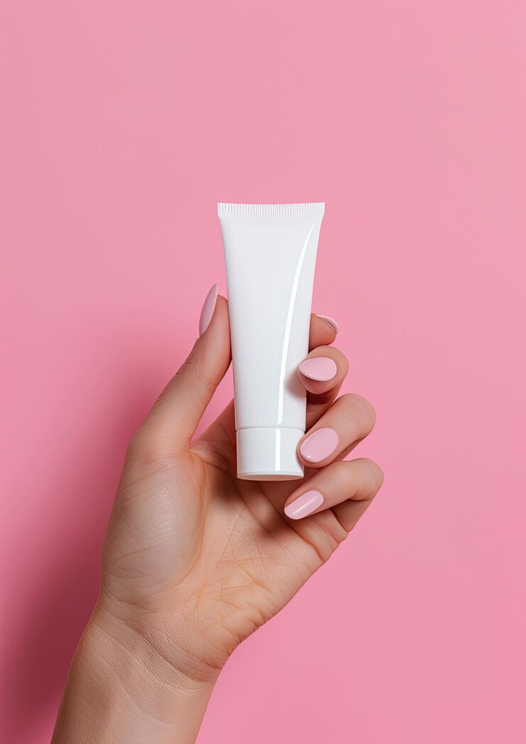 una mano sosteniendo un tubo de crema blanca contra un fondo rosa