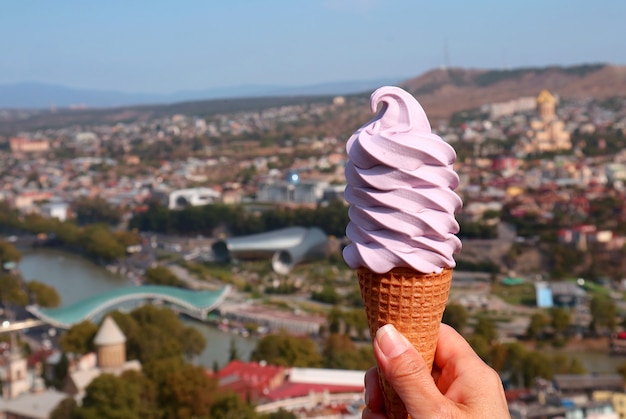 Mano sosteniendo un vino georgiano suave servir helado de cono contra borrosa pintoresca vista de la ciudad de Tbilisi