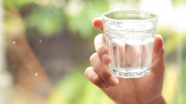 La mano sostiene el agua clara en un vaso. mano que sostiene un vaso transparente de agua mineral limpia. Concepto de protección del medio ambiente, bebida saludable. naturaleza fondo borroso