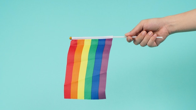 La mano sostiene una bandera del arco iris sobre fondo verde menta o azul tiffany.