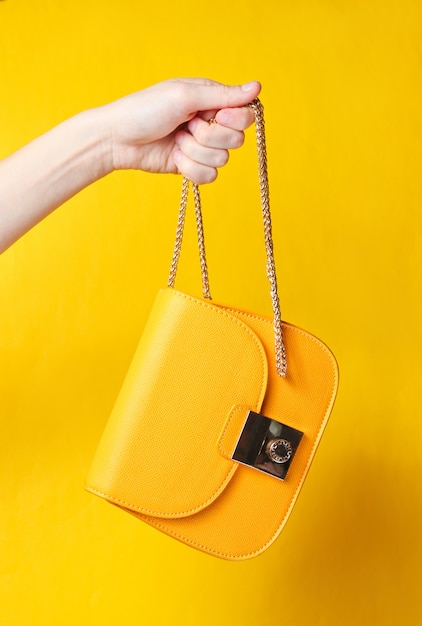 La mano sostiene el bolso de cuero amarillo de moda con cadena dorada sobre superficie amarilla