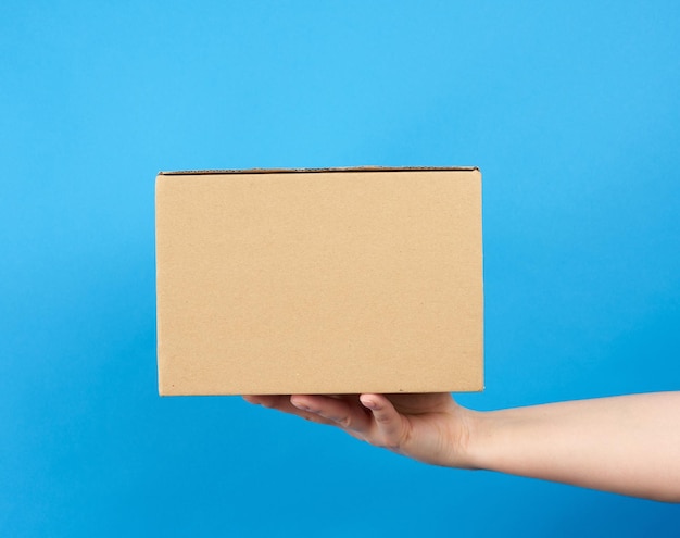 La mano sostiene una caja de cartón marrón de papel sobre un fondo azul de cerca