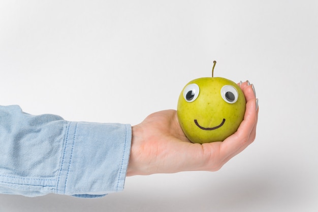 Foto la mano sostiene una carita verde de manzana con ojos saltones. apple divertido sobre fondo blanco