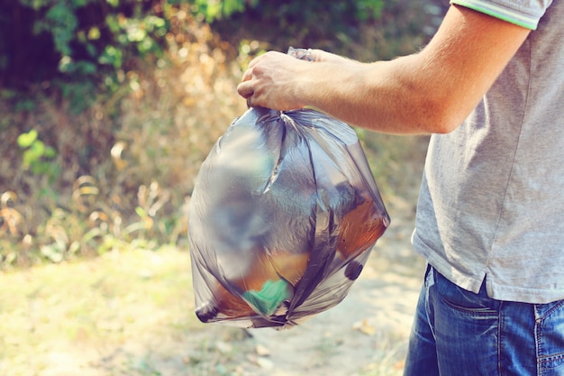 Una mano sostiene contra un bosque lleno de basura una gran bolsa de plástico negra, un día de verano