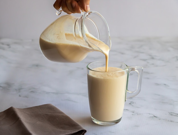 La mano sostiene una jarra de vidrio y vierte leche horneada fermentada en un vaso.