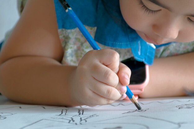 La mano sostiene un lápiz para dibujar en una lección de arte en papel, una niña adorable que sonríe alegremente mientras disfruta de una lección de arte y artesanía en el trabajo escolar