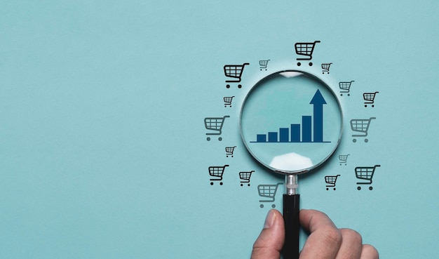 Una mano sostiene una lupa centrándose en un gráfico de barras azul con una tendencia ascendente rodeado de iconos de carrito en un fondo azul claro Esto simboliza el crecimiento del comercio electrónico y el concepto de análisis de mercado