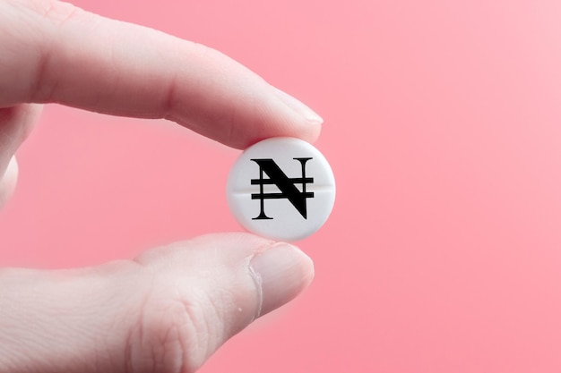 Una mano sostiene una píldora médica con un signo de moneda naira sobre un fondo rosa El concepto de un aumento en el precio de los medicamentos en Nigeria