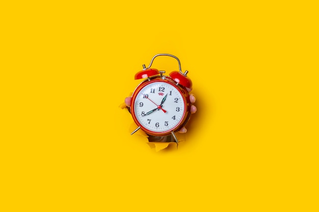 La mano sostiene un reloj rojo en un agujero en un fondo amarillo roto.