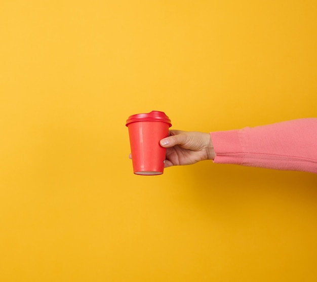 La mano sostiene una taza roja de cartón de papel para café, fondo amarillo. vajilla ecologica
