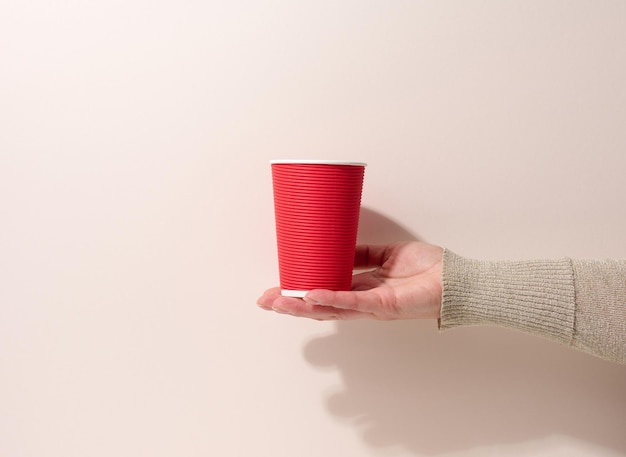 La mano sostiene la taza roja de cartón de papel para café, fondo beige. Vajilla ecológica, cero residuos