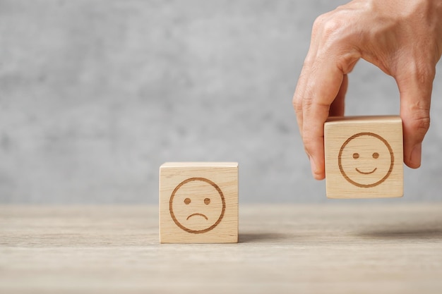 Mano sujetando el bloque de cara de sonrisa. El cliente elige Emoticon para las reseñas de los usuarios. Calificación del servicio, clasificación, revisión del cliente, satisfacción, estado de ánimo, salud mental y concepto de retroalimentación